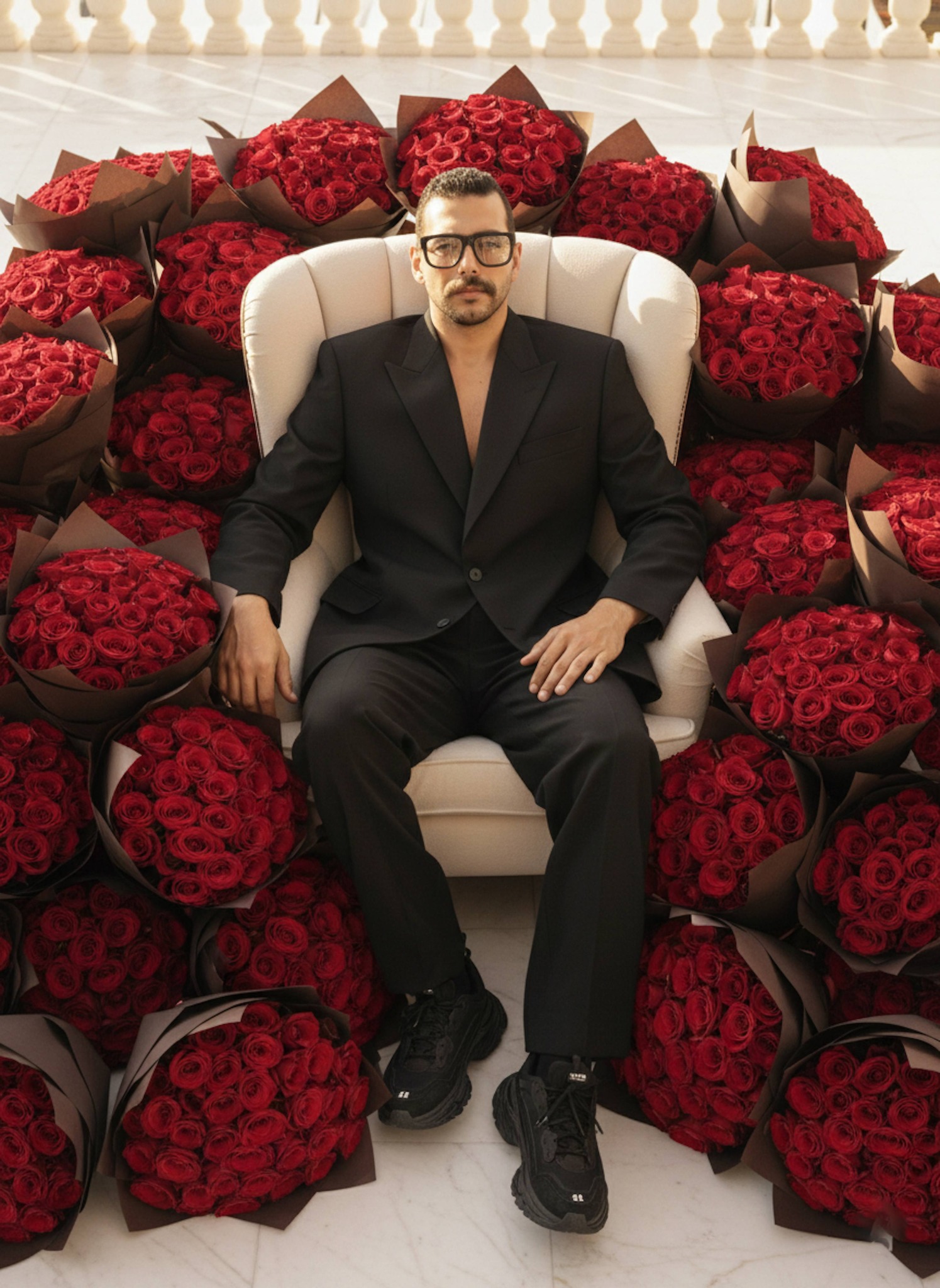 Person in elegant suit seated in cream armchair surrounded by massive rose bouquets