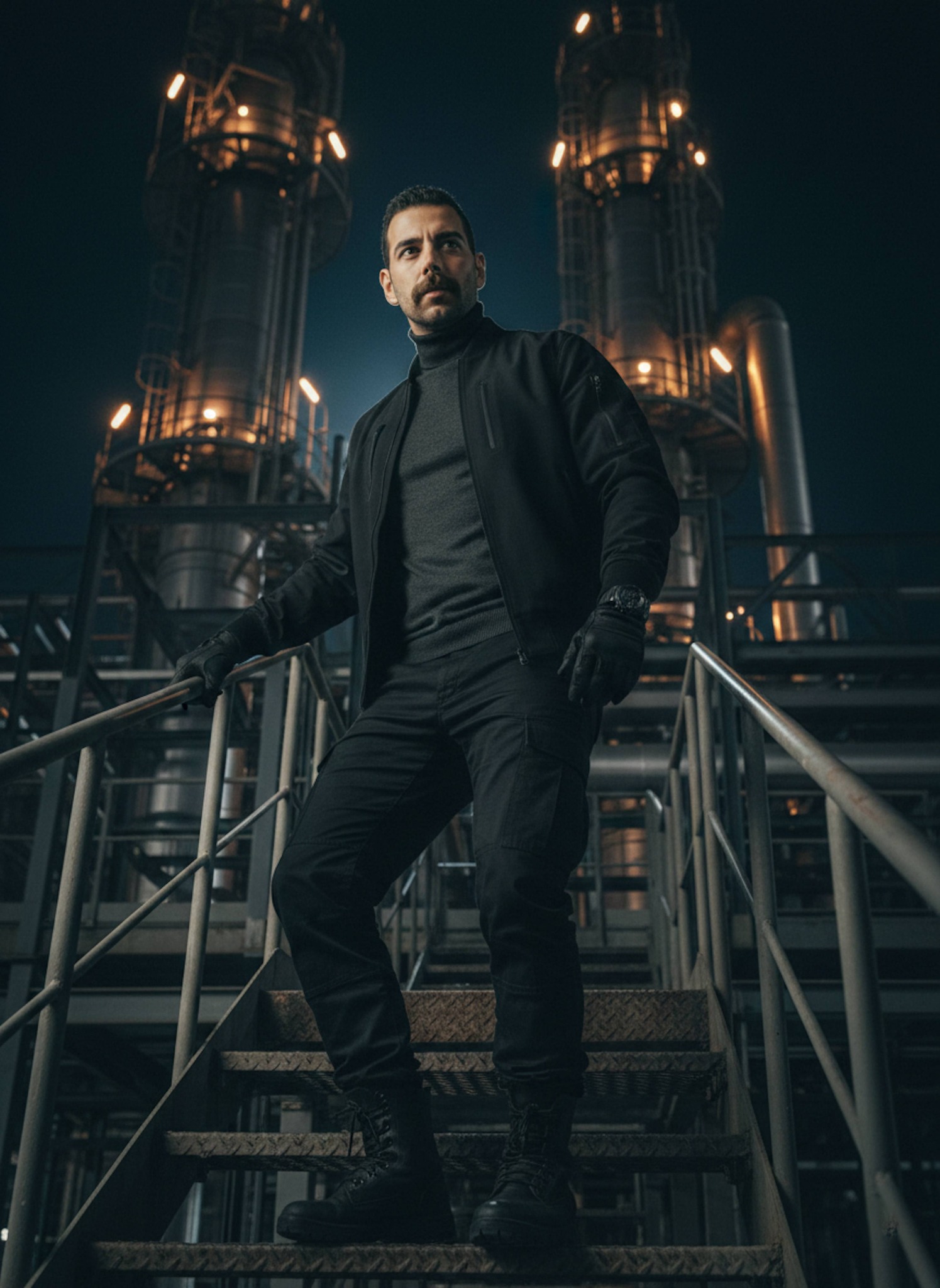 Person in black bomber jacket on refinery metal stairs at night with orange warning lights and pipes
