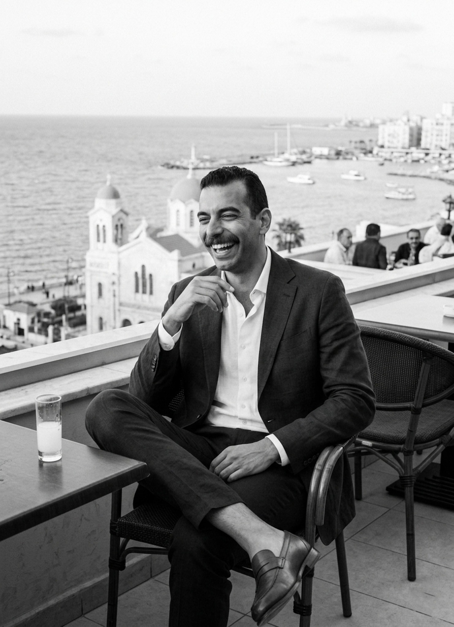 Person laughing in a tailored linen blazer at the Greek Club with the Mediterranean Sea visible behind in elegant monochrome