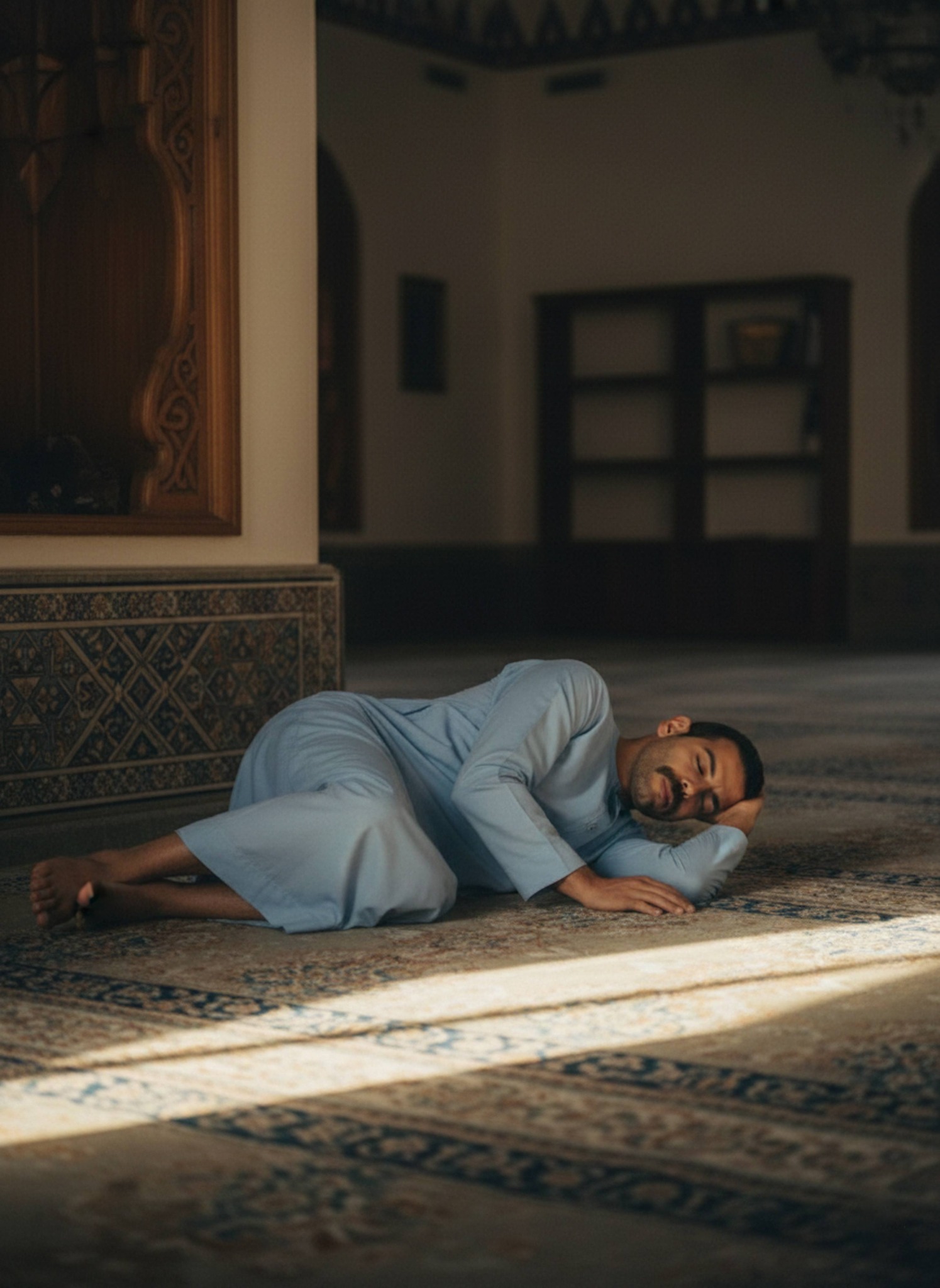 Person in light blue galabeya napping on mosque carpet lying on side with hand under cheek and sunlight ray nearby