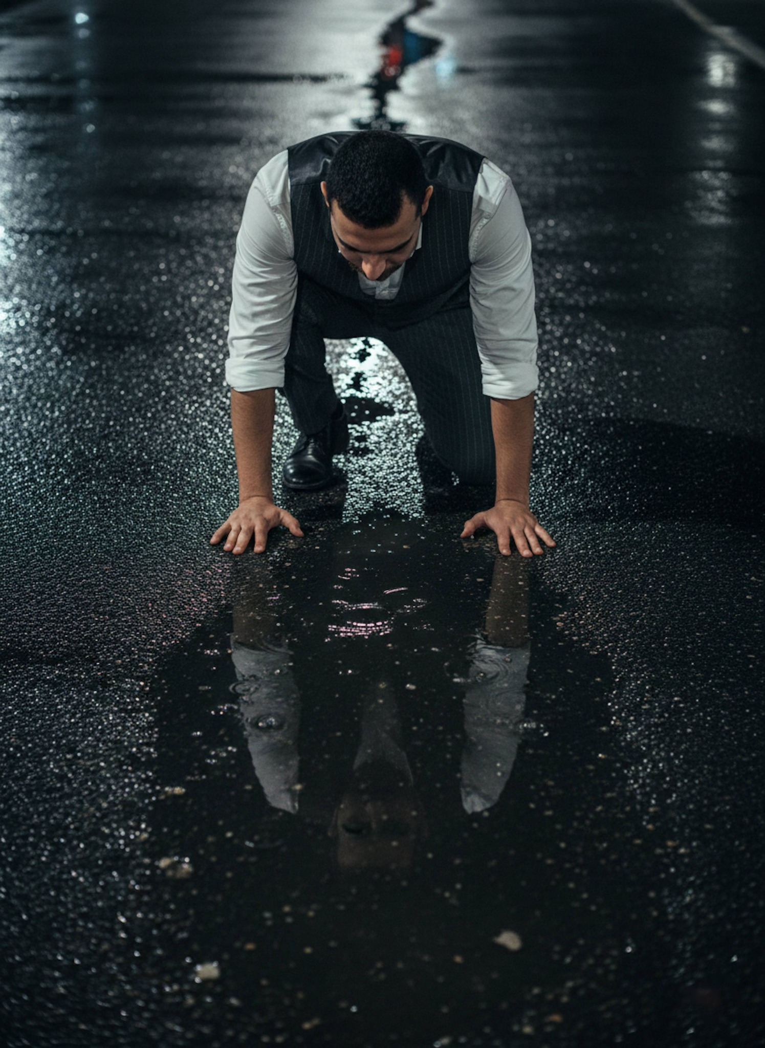 Person in charcoal pinstripe vest kneeling to look at reflection in rain puddle on dark paved road at night