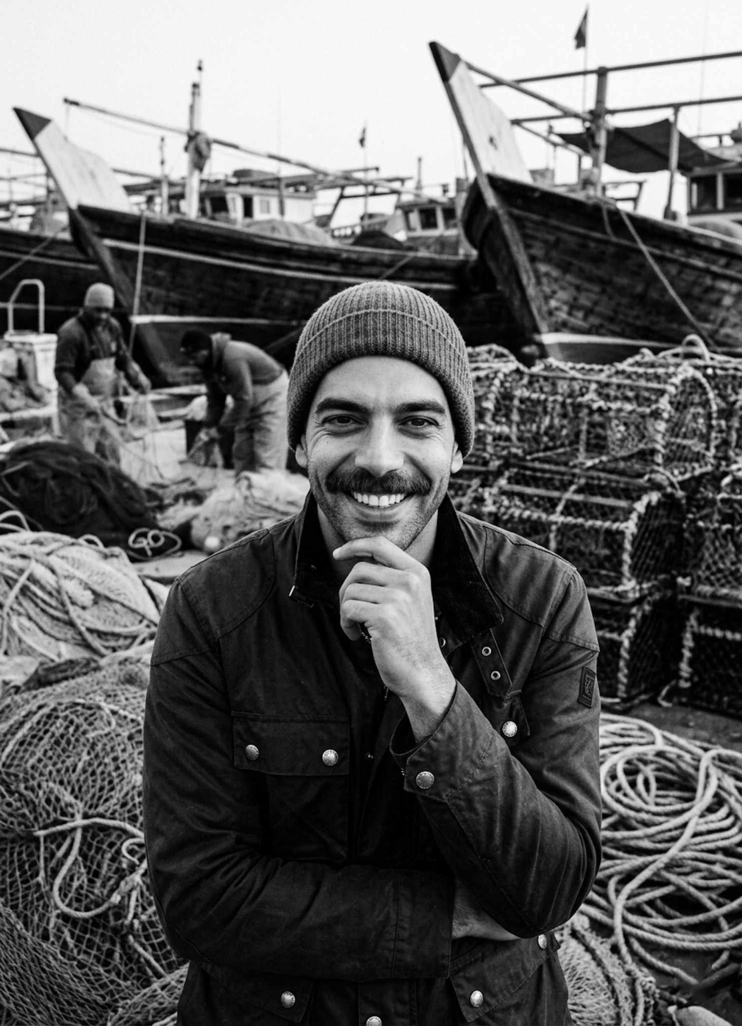 Person in waxed canvas field jacket and beanie at Bahary fishing port with fishing nets and wooden boats in monochrome