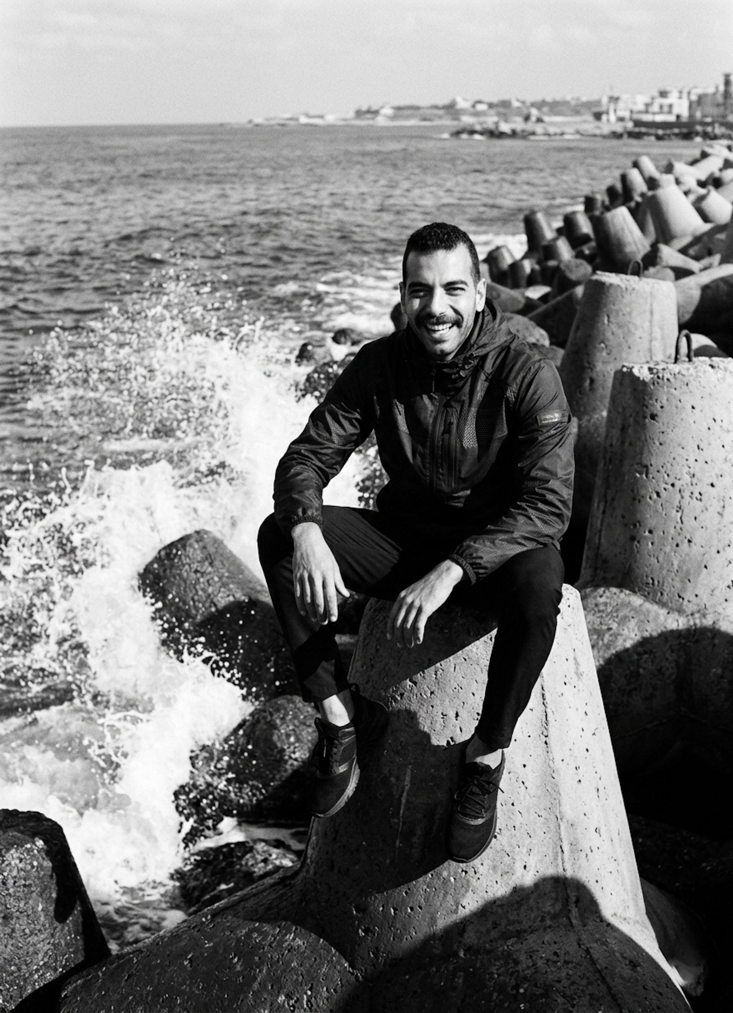 Person in designer windbreaker and modern sneakers at Gleem Bay with sea waves crashing on concrete tetrapods in monochrome