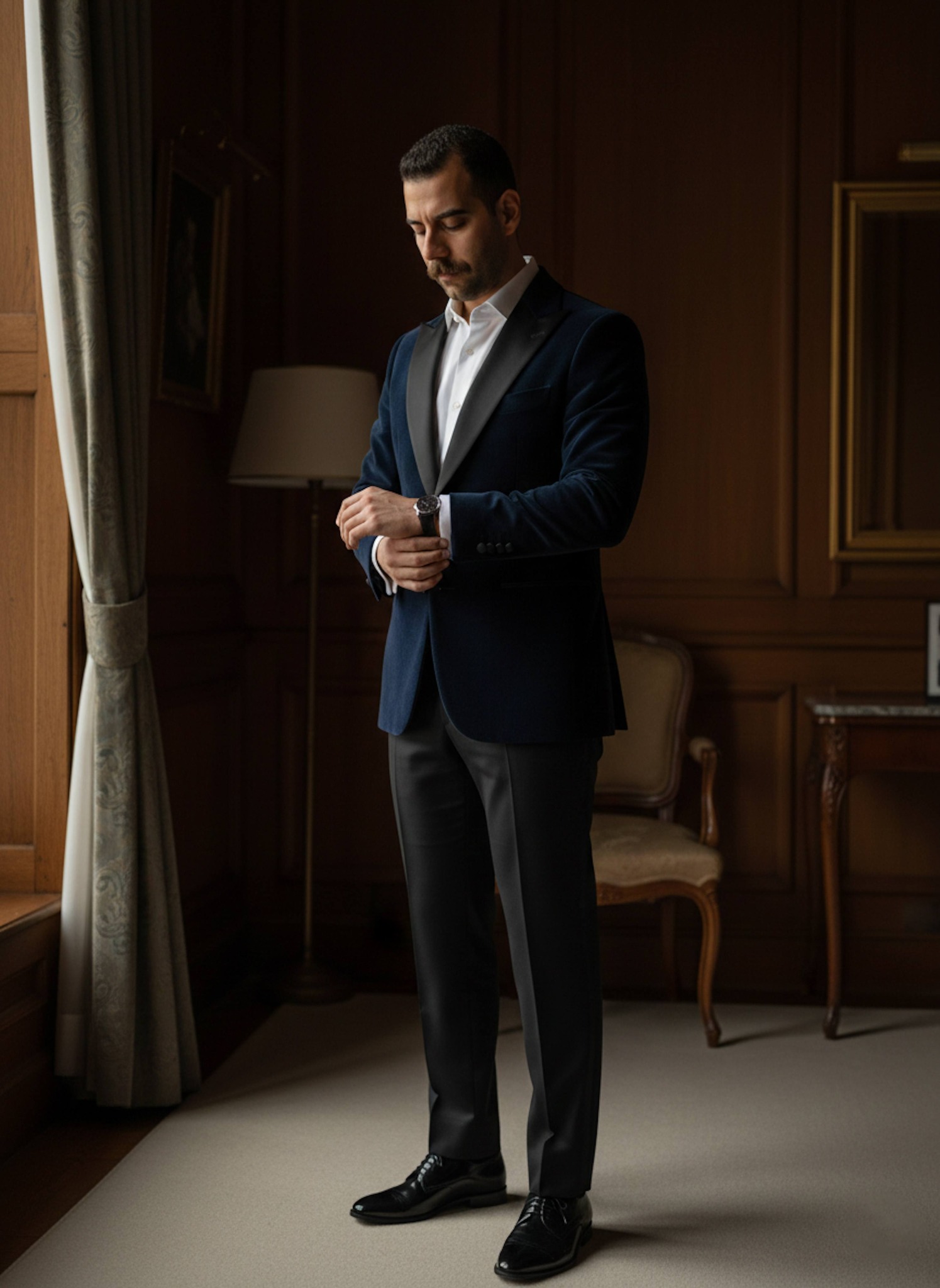 Groom in midnight-blue velvet tuxedo standing in a dimly lit wood-paneled historic hotel suite