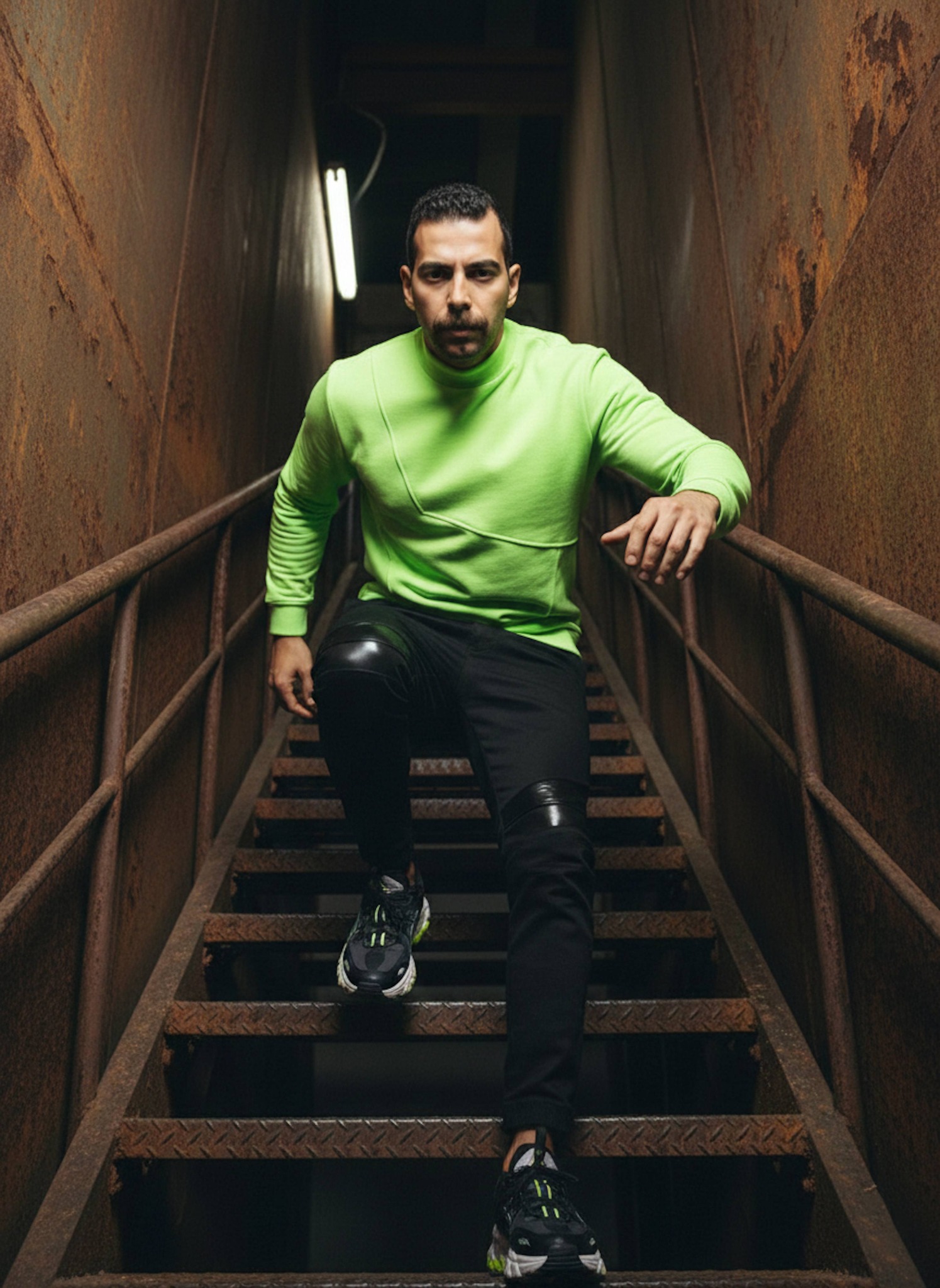 Person in neon green sweatshirt posing in a narrow rusted industrial stairwell with harsh lighting