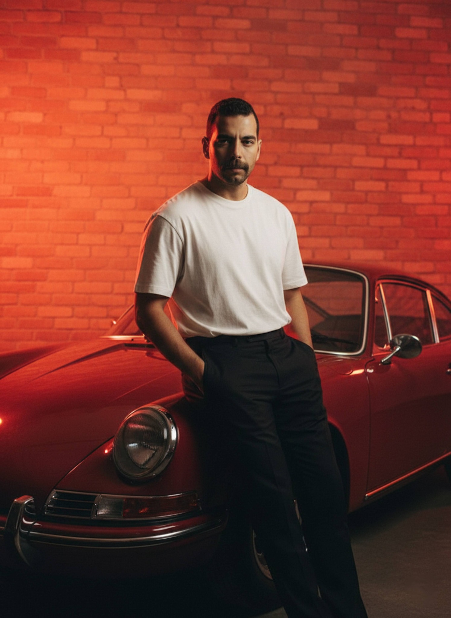 Cinematic portrait of a person leaning against a red 1960s vintage sports car with dramatic orange backlighting